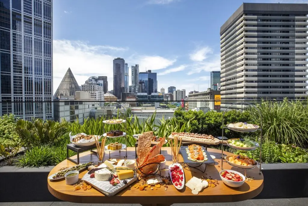 Outdoor table with cheese, fruit and assorted dishes surrounded by greenery with Melbourne city views