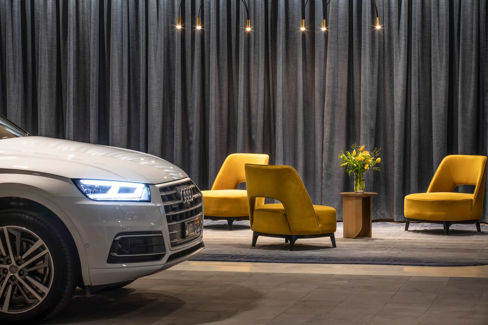 Silver car beside mustard lounge chairs, pendant lights and floor-to-ceiling grey curtains