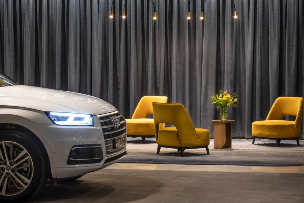 Silver car beside mustard lounge chairs, pendant lights and floor-to-ceiling grey curtains
