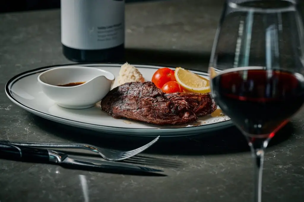 Grilled steak served with tomatoes, lemon and sauce beside a glass of red wine