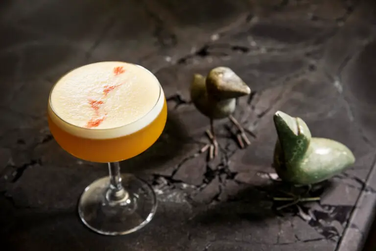 Foamy golden cocktail in a coupe glass beside two small bird sculptures on a dark surface