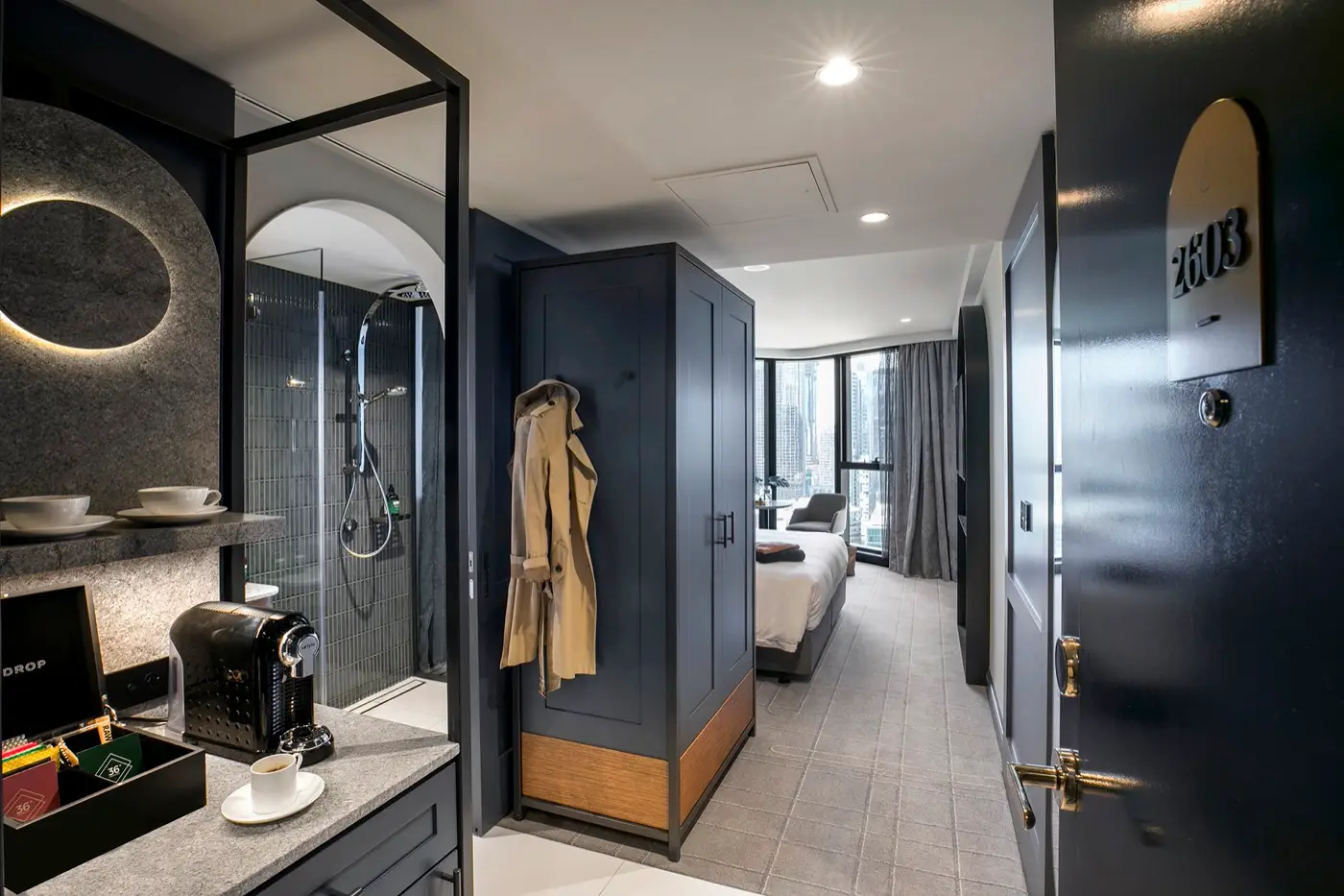 Guest room entrance with coffee station, open bathroom, wardrobe and bed beside city-facing windows