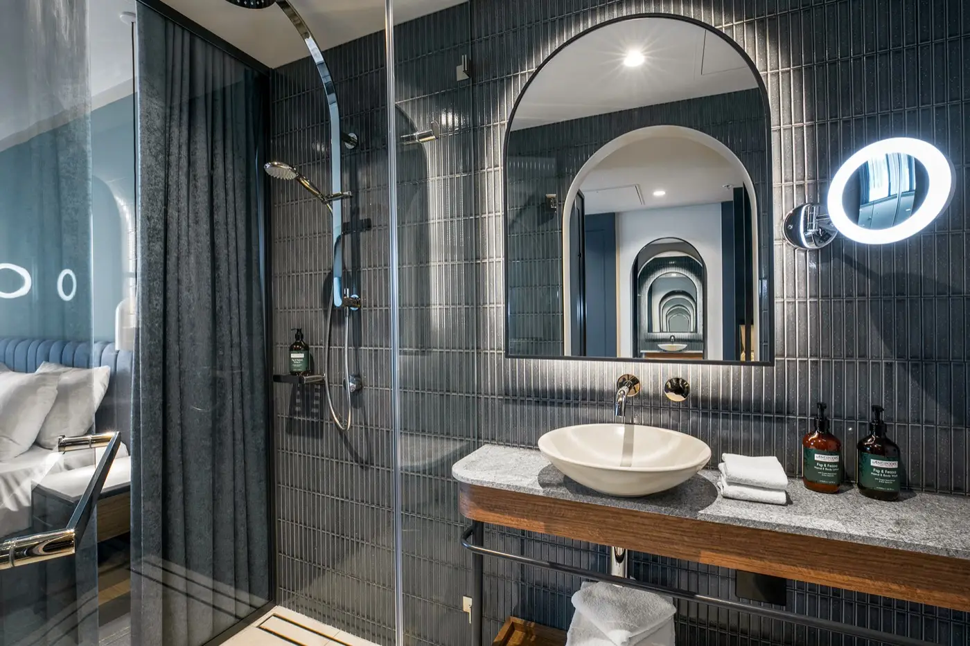 Bathroom with glass shower, basin, arched mirror and dark tiled walls