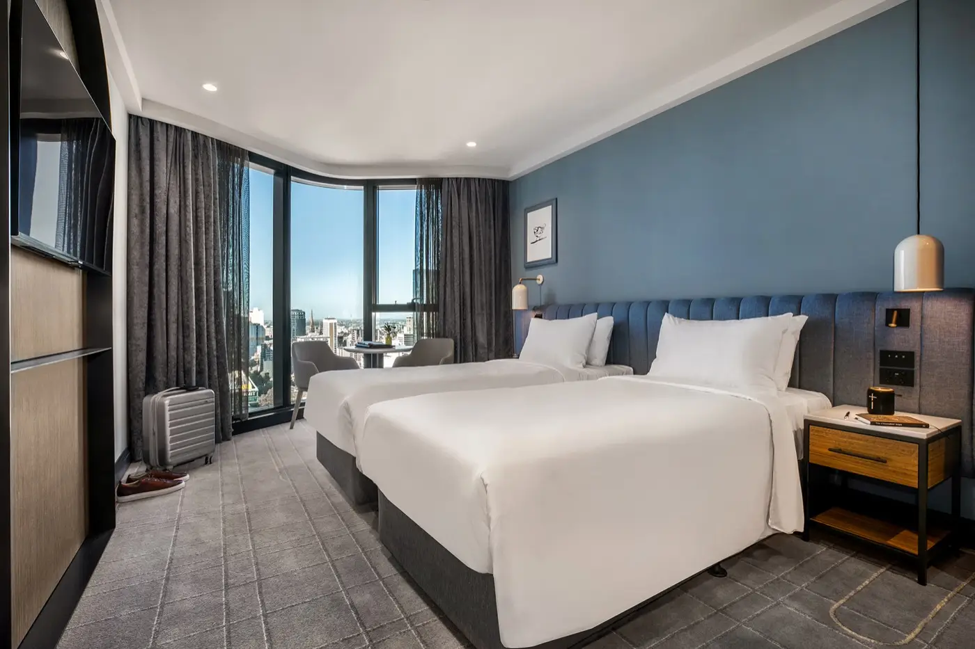 Guest room with two beds, blue upholstered headboard and floor-to-ceiling windows overlooking Melbourne