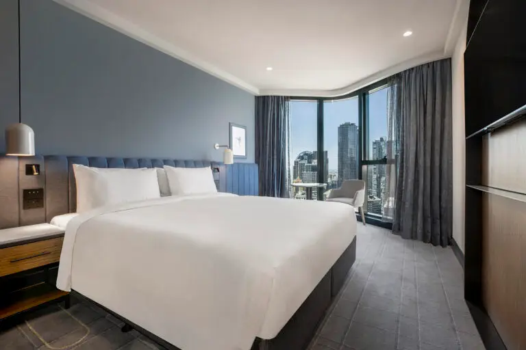 Guest room with two beds, blue upholstered headboard and floor-to-ceiling windows overlooking Melbourne