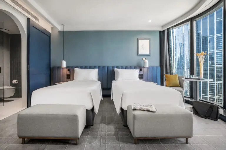 Twin guest room with two beds, upholstered benches and floor-to-ceiling windows overlooking the city