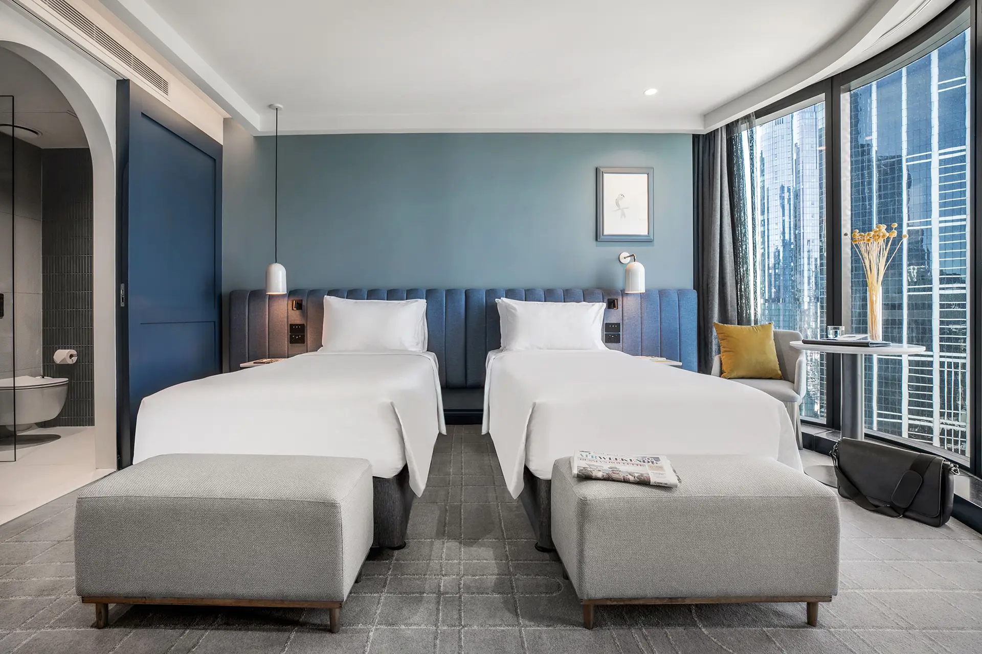 Twin guest room with two beds, upholstered benches and floor-to-ceiling windows overlooking the city