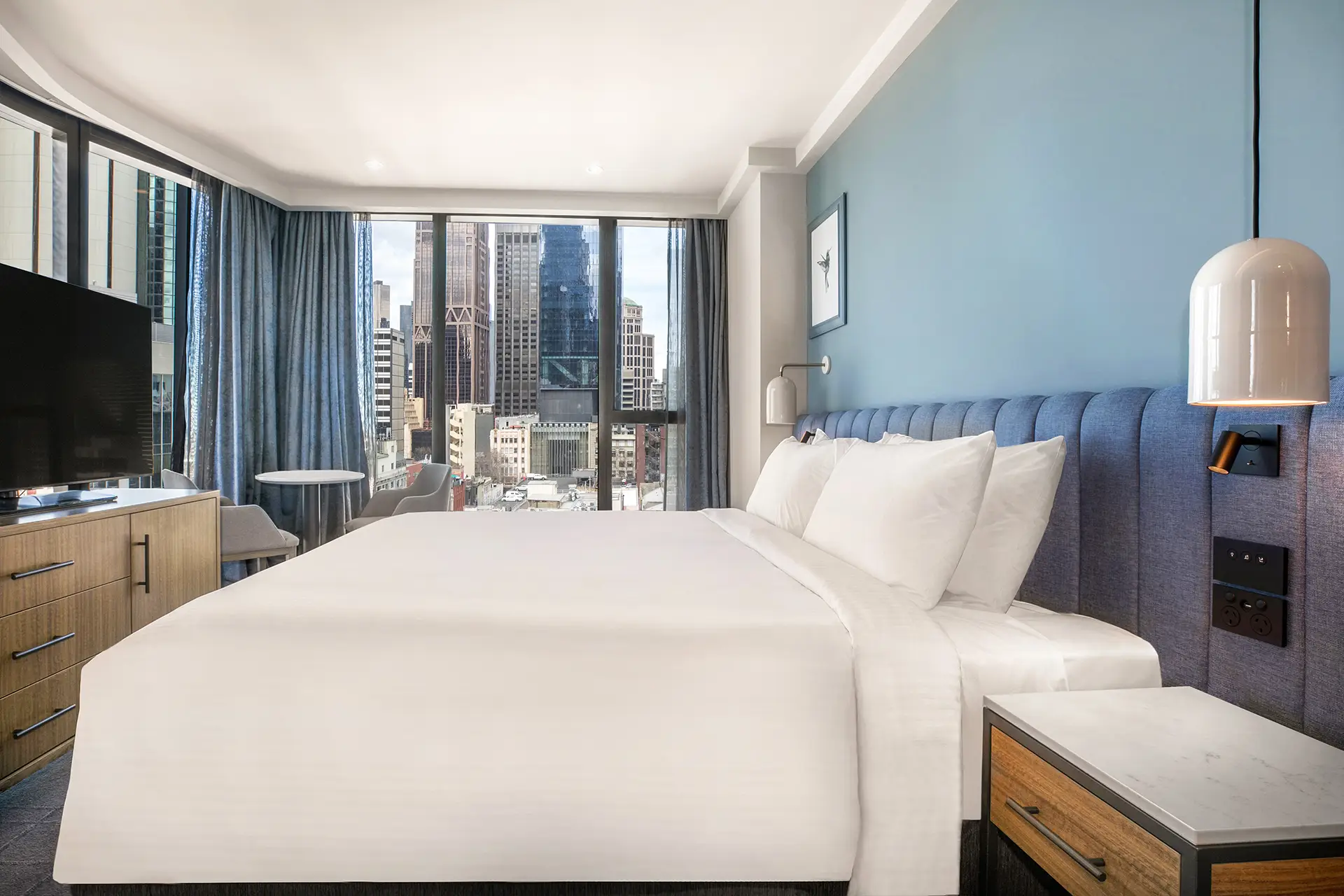 King guest room with blue upholstered headboard, window-side seating and views across city buildings