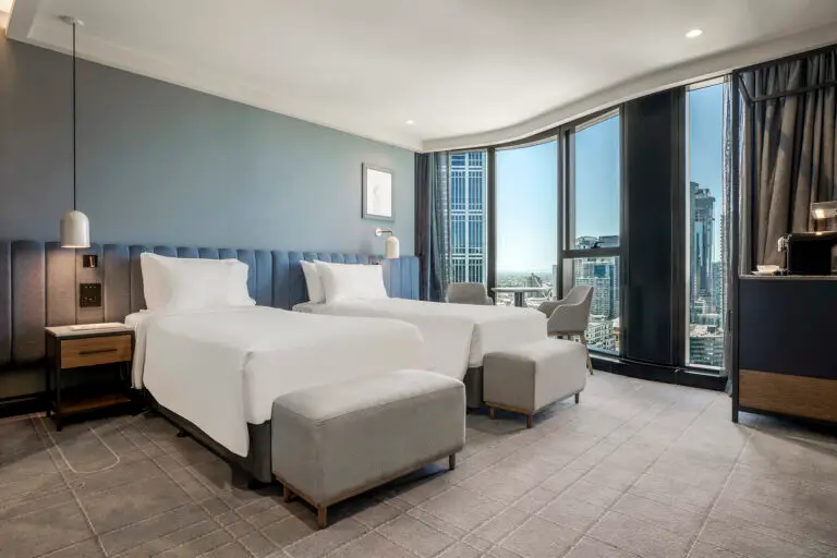 Twin guest room with two beds, upholstered benches and curved floor-to-ceiling windows overlooking the city