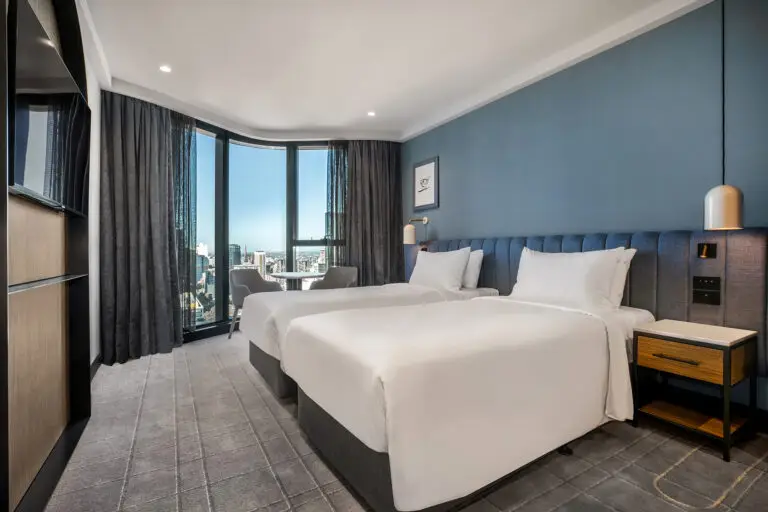 Guest room with two beds, blue upholstered headboard and floor-to-ceiling windows overlooking Melbourne