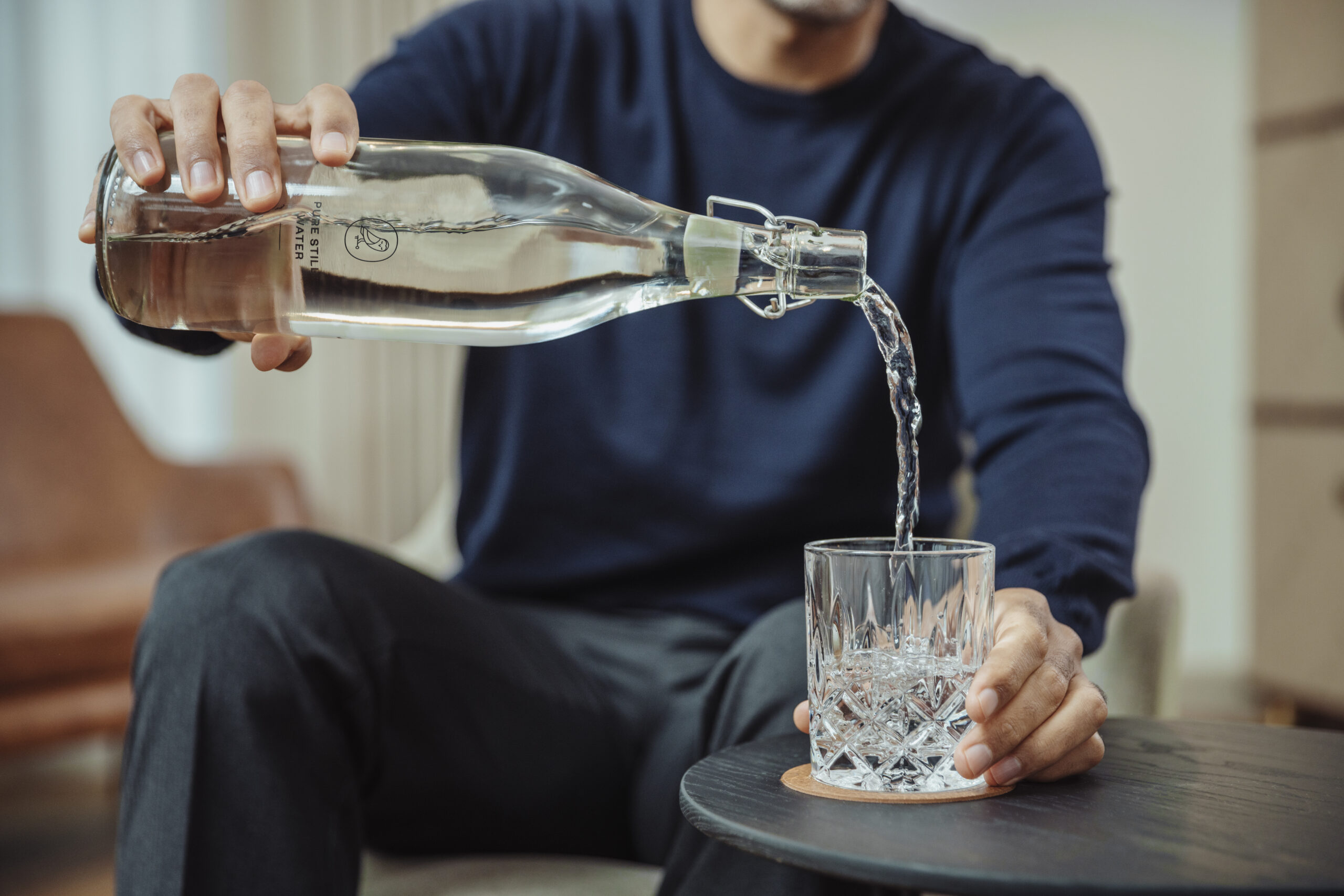 Guest pouring water from a glass bottle into a crystal tumbler on a side table in a hotel room