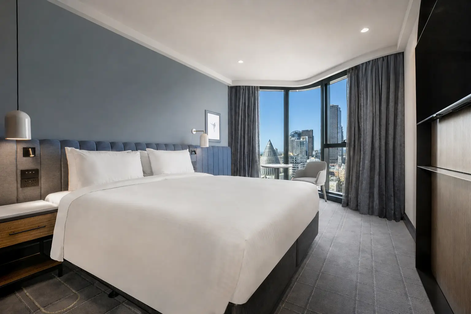 Guest room with a large bed, blue upholstered headboard and curved windows overlooking Melbourne
