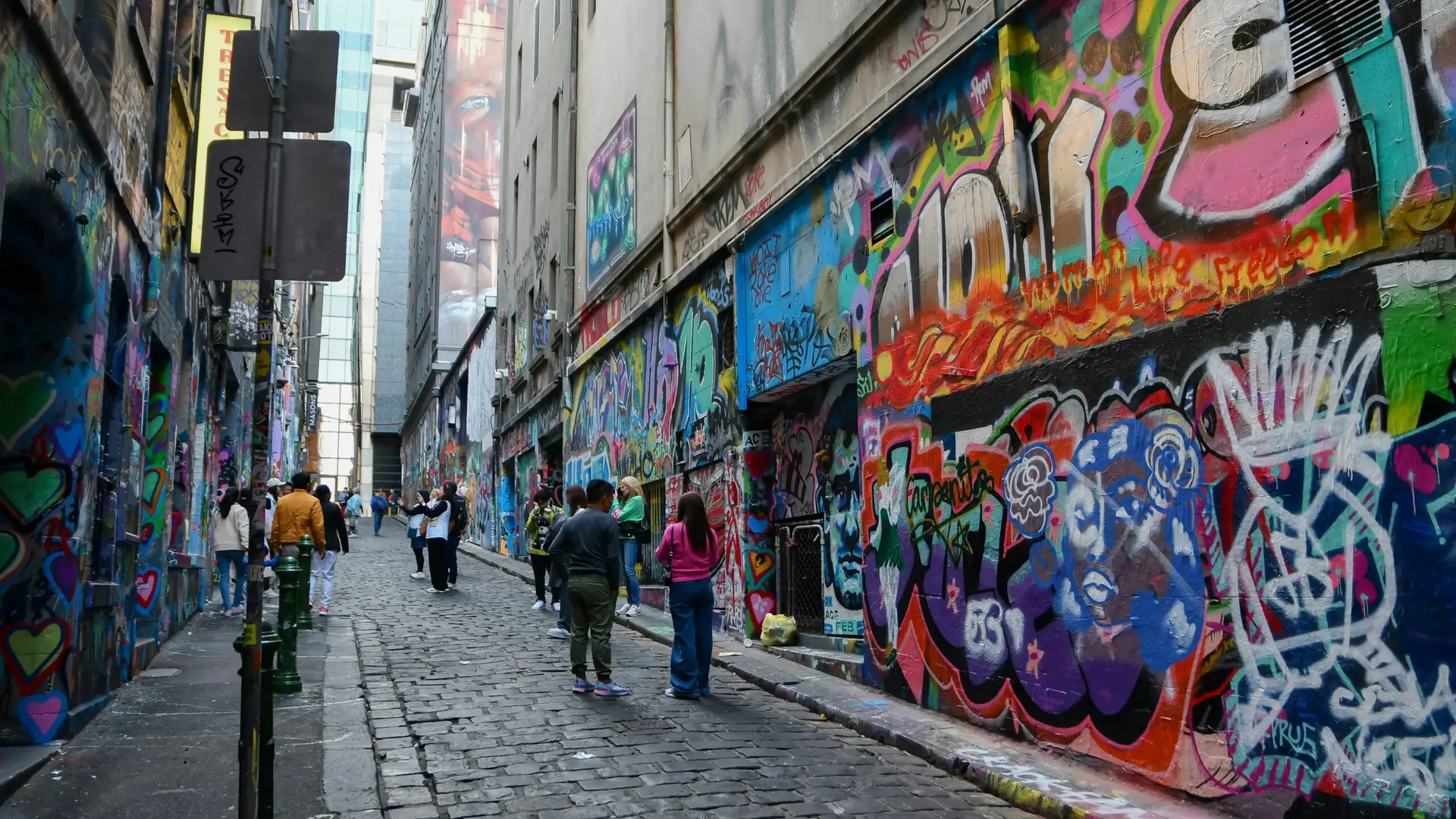 People walking down a cobblestone laneway covered in colourful graffiti and street art murals on brick walls.