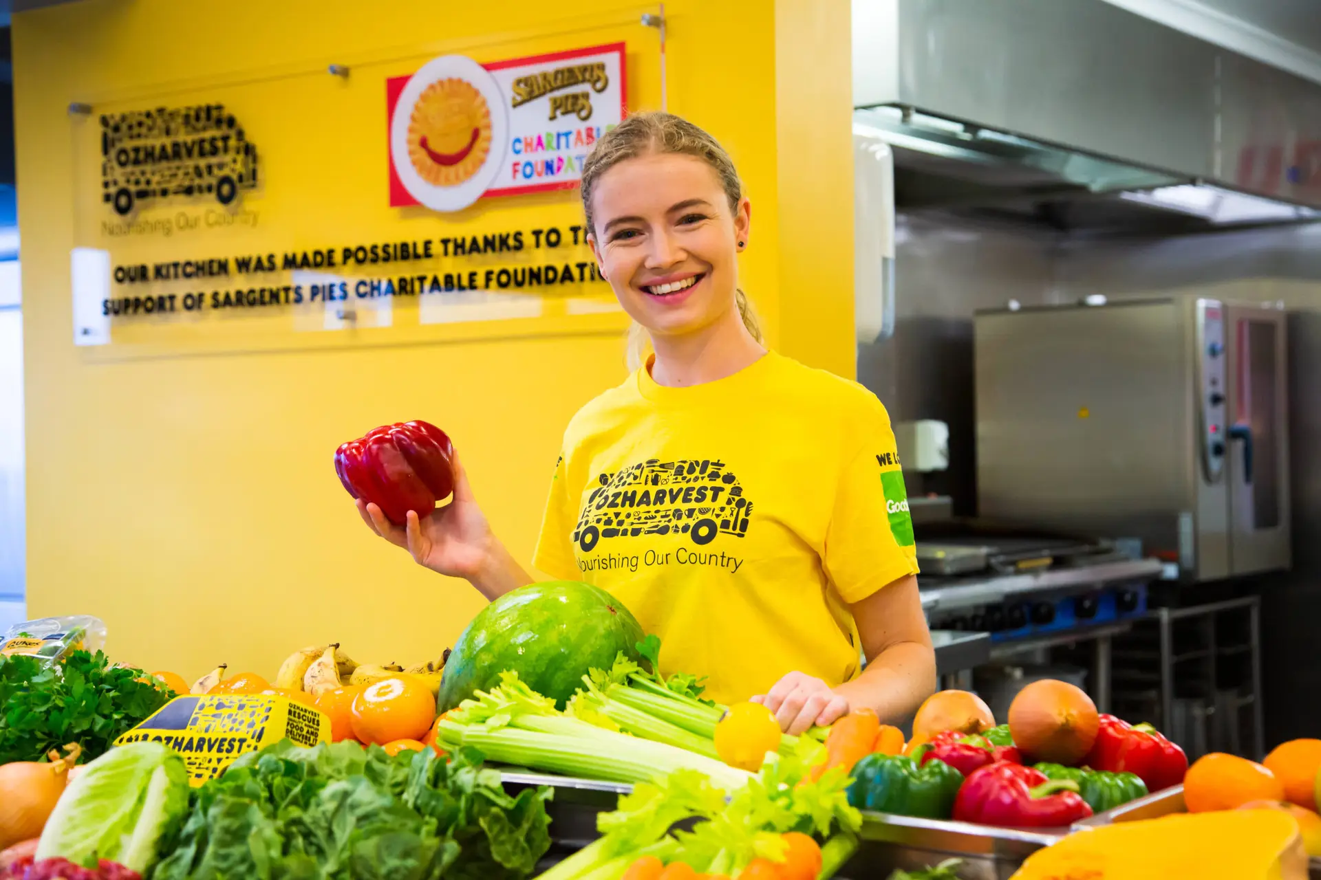 Smiling team member in an OzHarvest shirt standing behind fresh fruit and vegetables