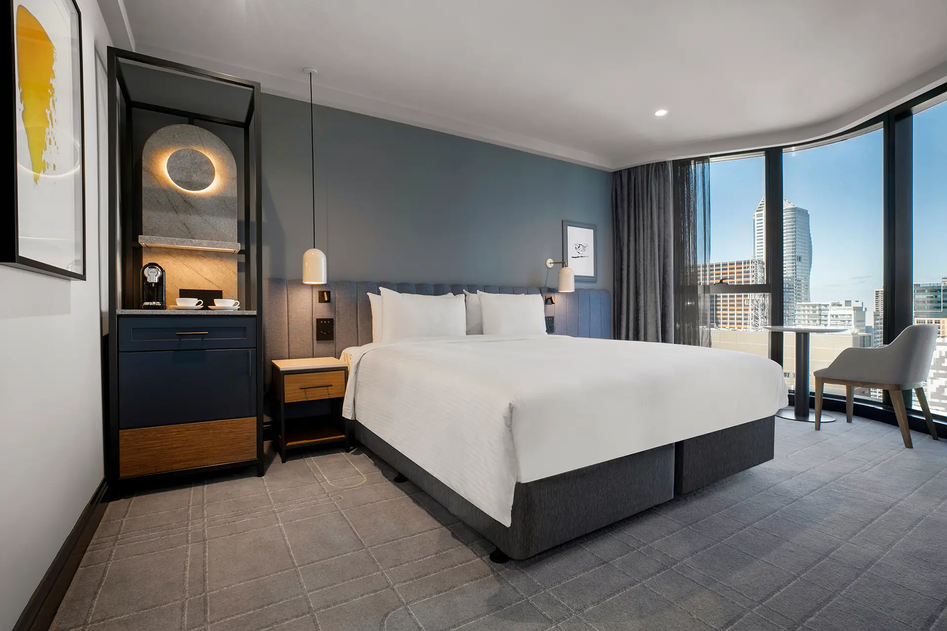 Guest room with white bed, blue upholstered headboard and curved windows overlooking Melbourne
