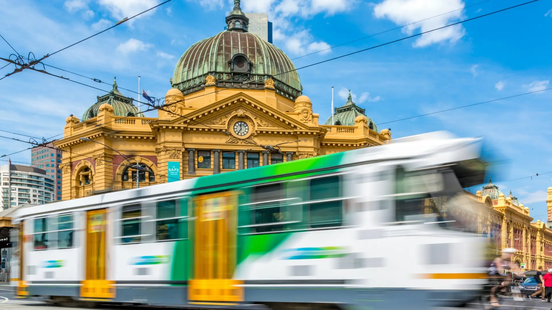 Modern green and white tram blurred in motion passing historical yellow building on city street.