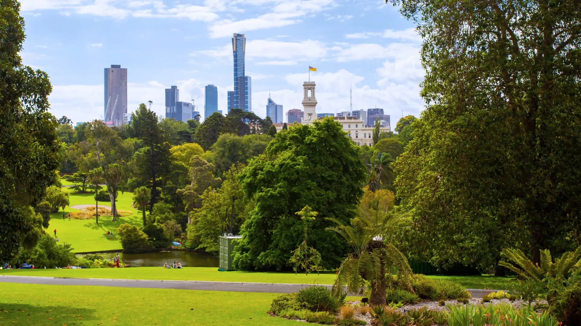 Lush green botanical gardens with tall trees and lawn overlooking the Melbourne city skyline.