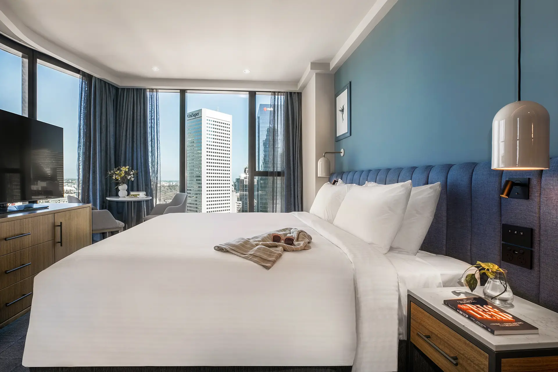 Guest room with white bed, blue headboard, timber cabinetry and windows overlooking the city