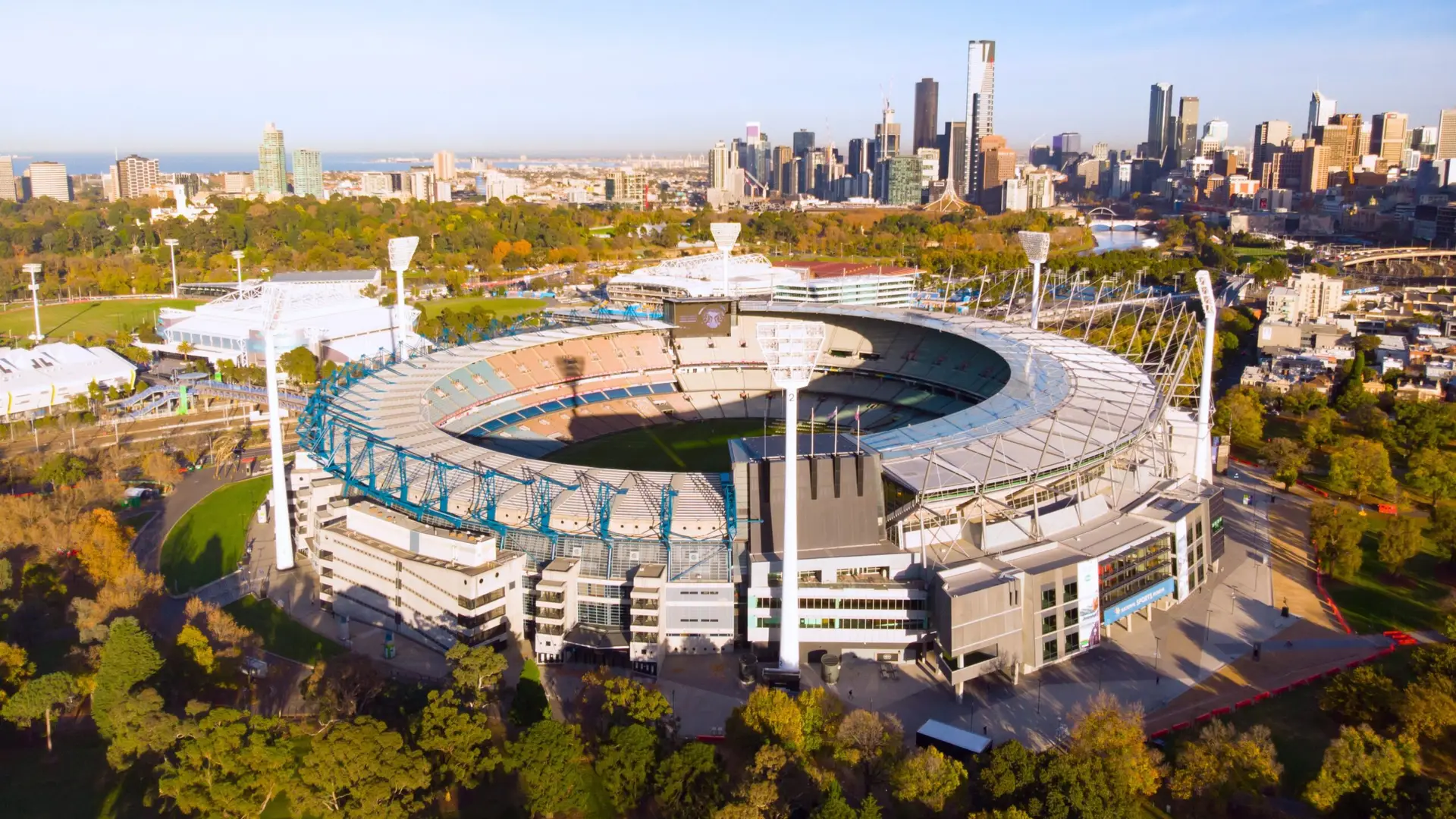 Aerial view of a large circular sports stadium surrounded by trees and urban cityscape.