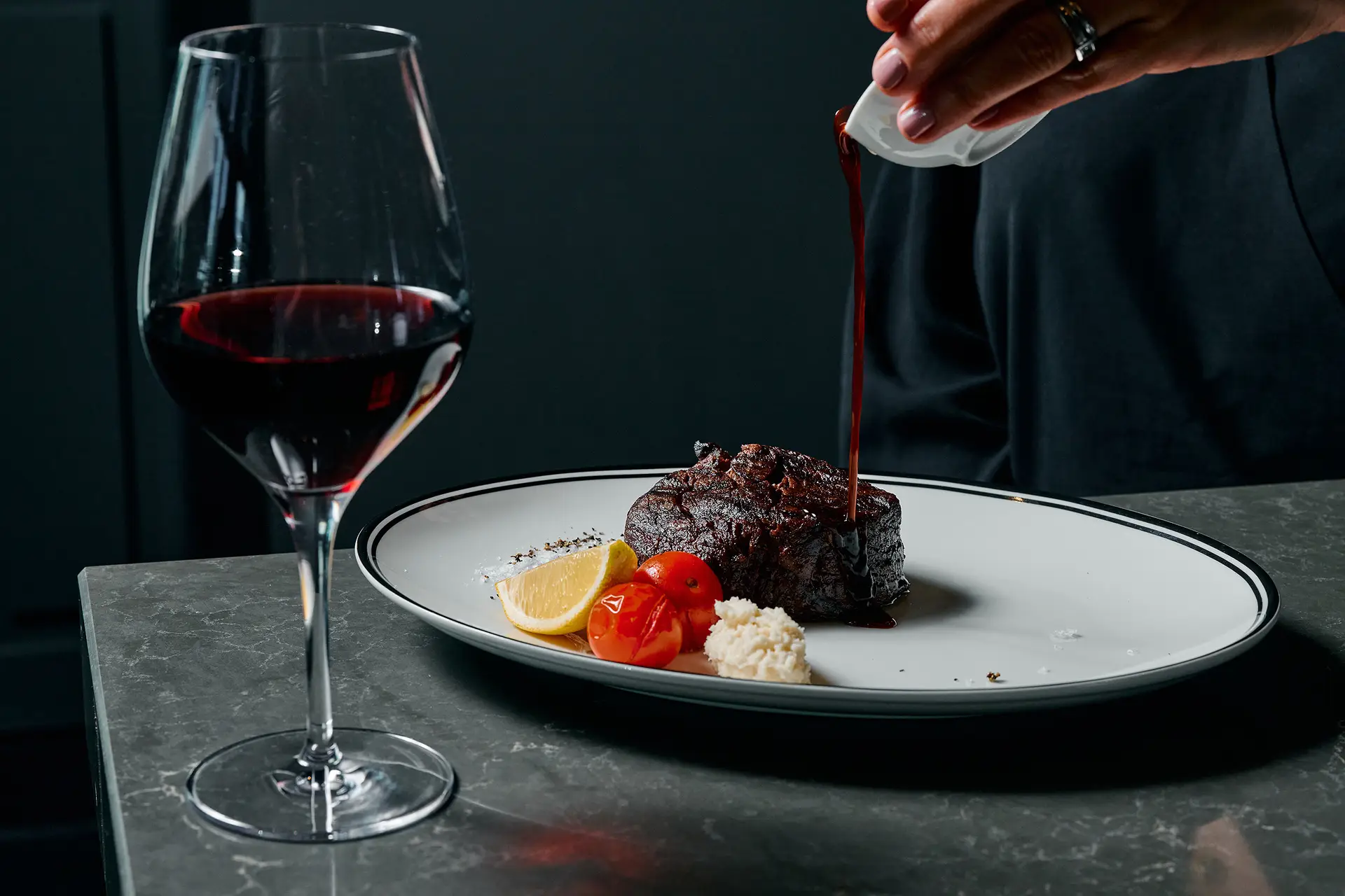 Plated steak with tomatoes and sauce being poured beside a glass of red wine