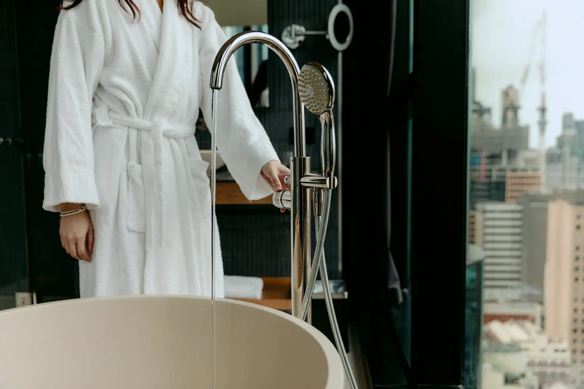 Guest in a white robe filling a freestanding bathtub beside a city-view window