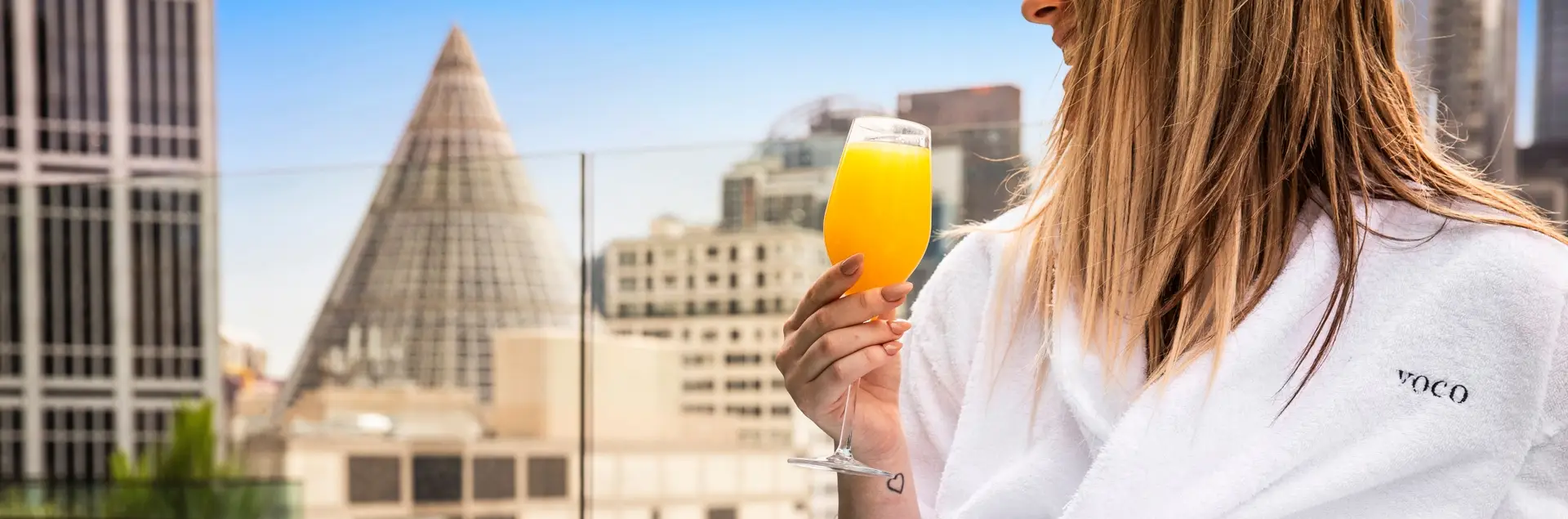 Guest in a white voco robe holding an orange drink with the Melbourne Central Shot Tower cone behind