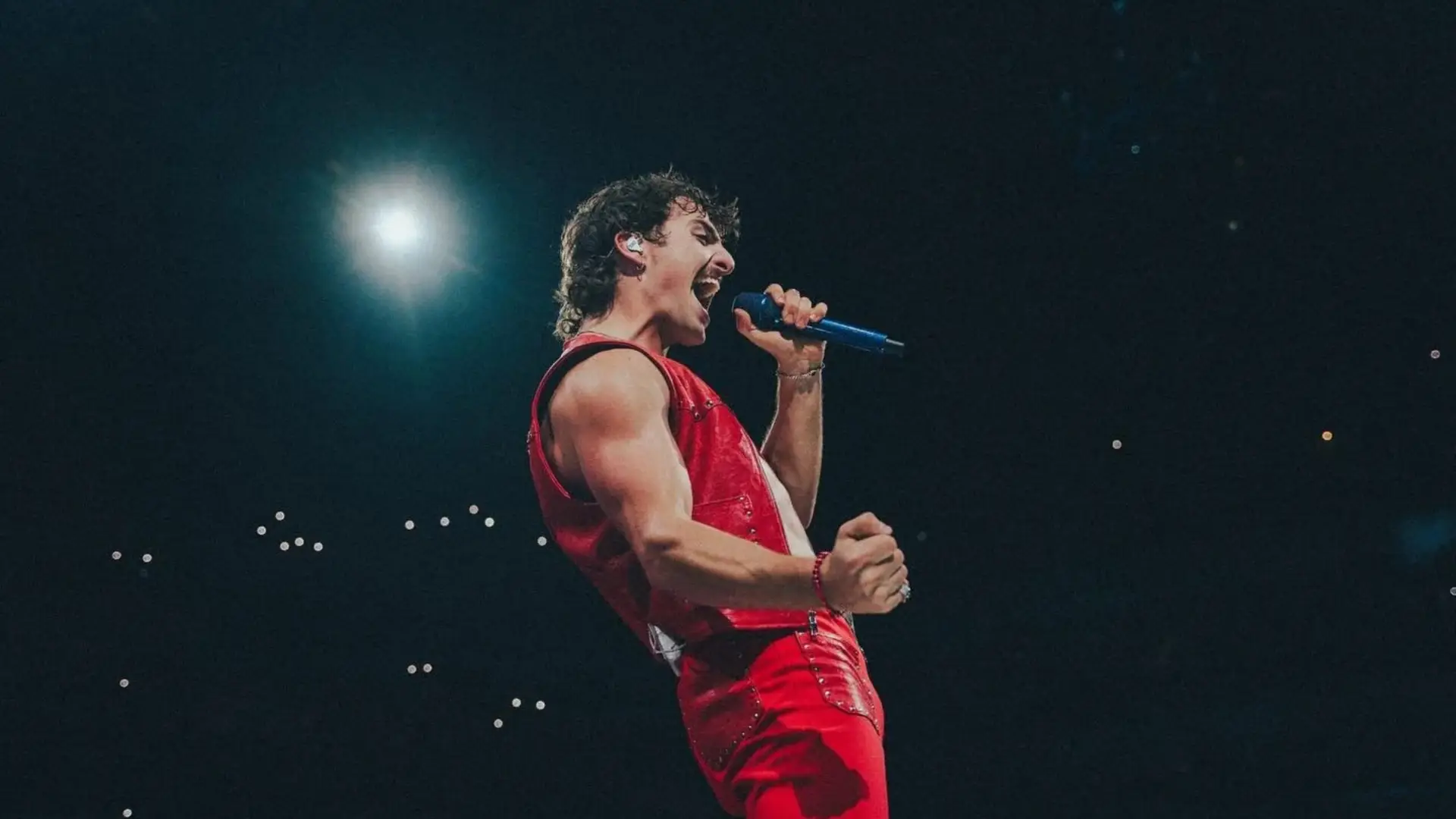 Male singer in a red leather vest and trousers performing on a dark stage while holding a blue microphone and singing into a light.