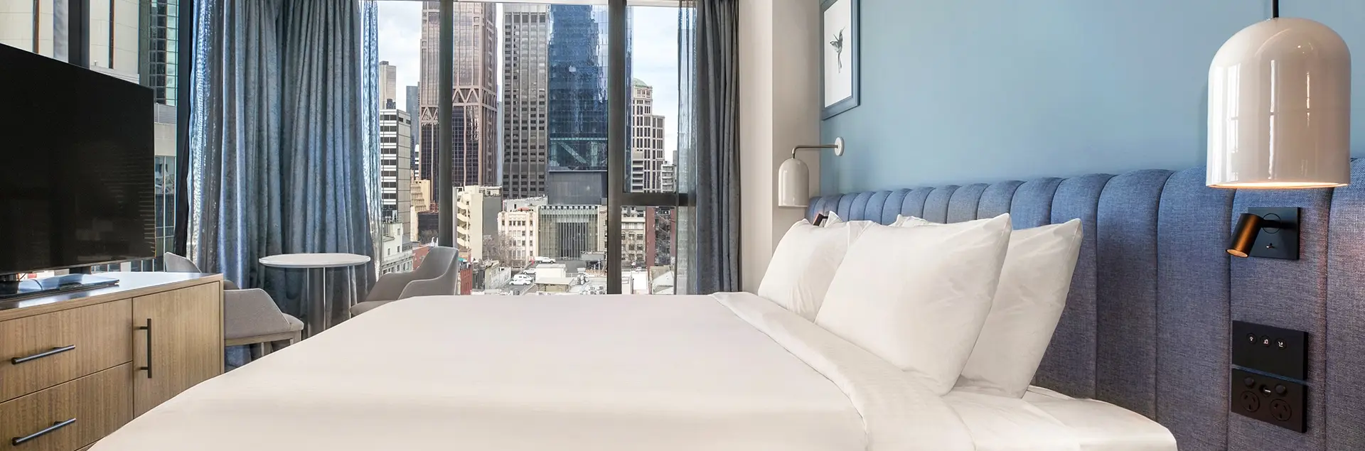 Guest room with a large bed, blue upholstered headboard and floor-to-ceiling windows overlooking the city