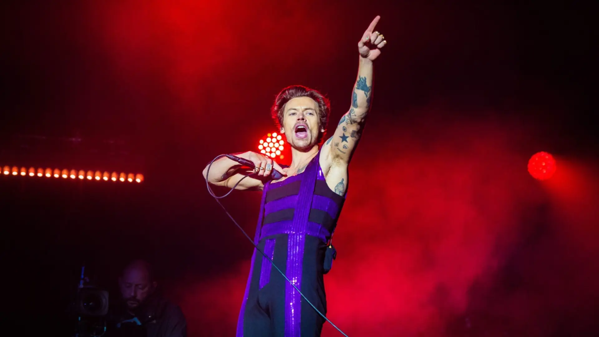 Male singer in a sparkly purple and black stage costume raising his arm and singing into a microphone under red concert lighting.