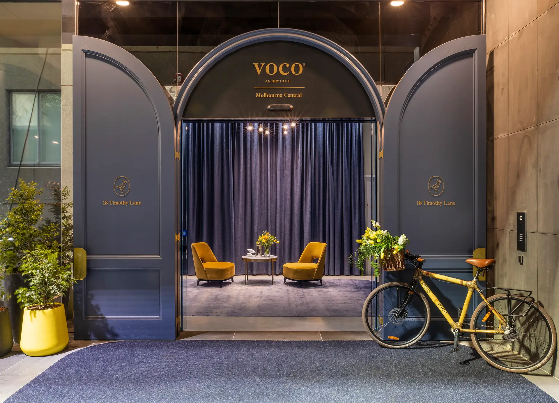 Arched Timothy Lane entrance with voco signage, mustard chairs, flowers and a bicycle