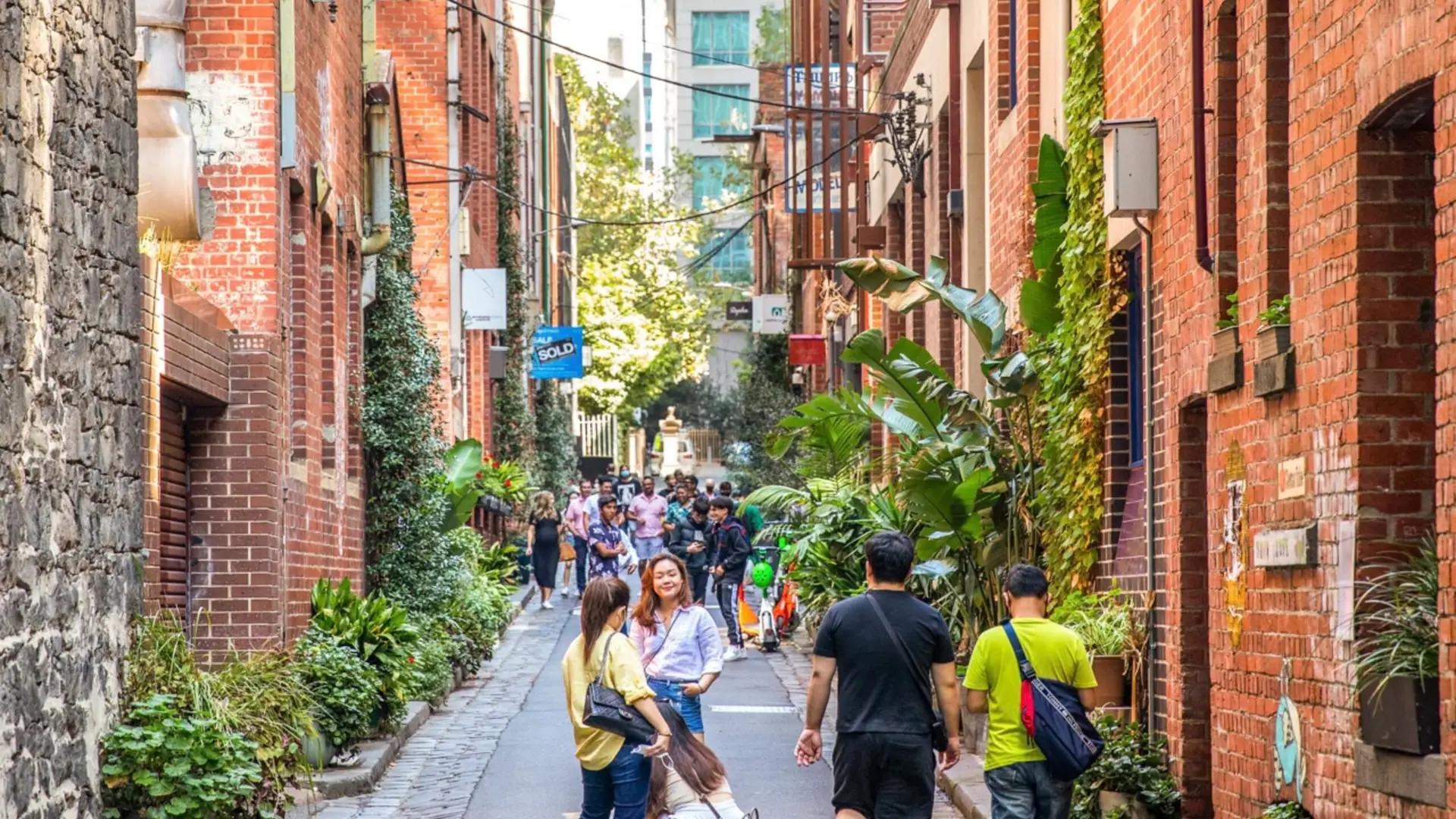 Pedestrians walking down a narrow urban laneway flanked by historic red brick buildings and green foliage.