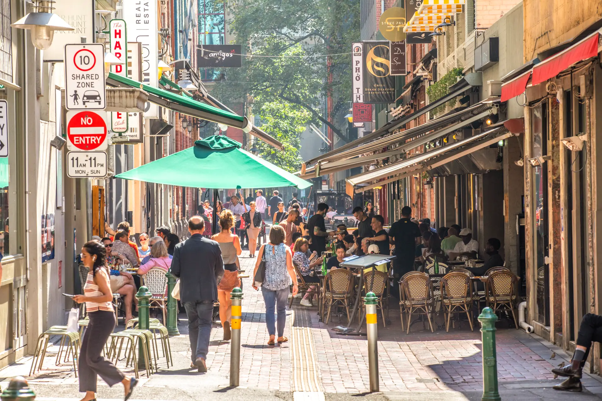 People dining at outdoor tables under a green sun umbrella in a busy urban alleyway lined with restaurant signs.