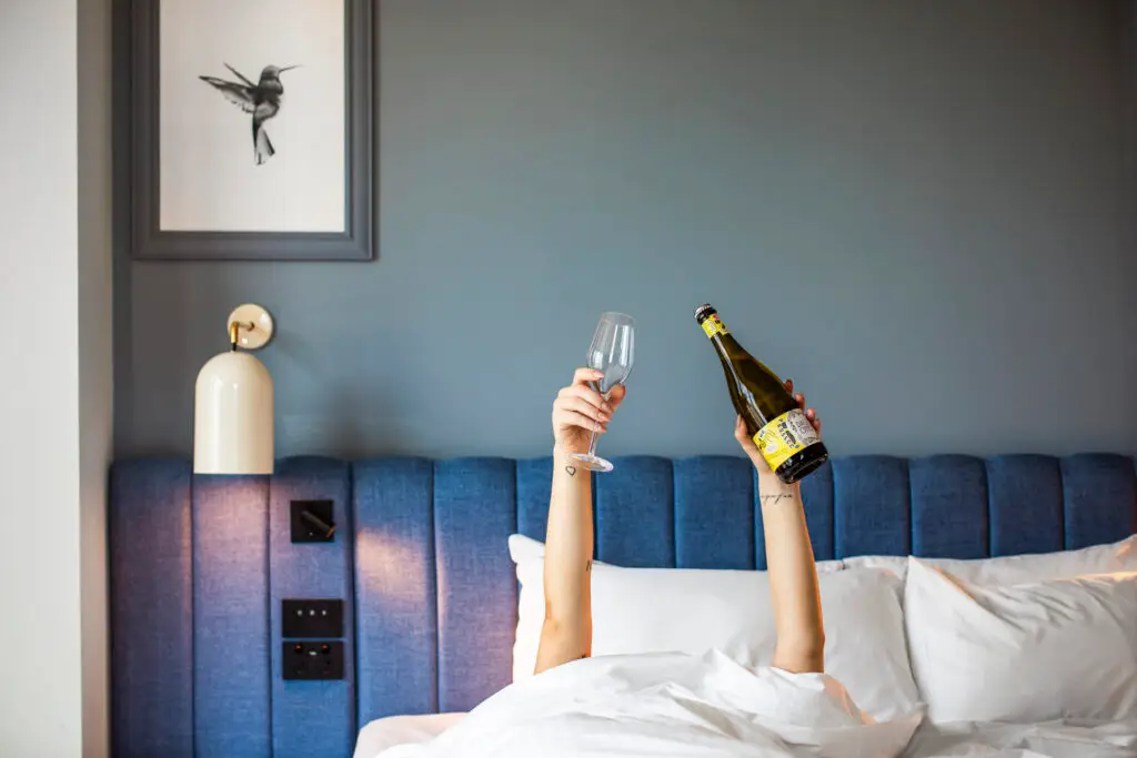 Guest in bed holding a bottle of prosecco and an empty glass against a blue upholstered headboard