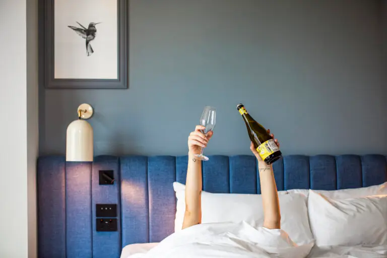 Guest in bed holding a bottle of prosecco and an empty glass against a blue upholstered headboard