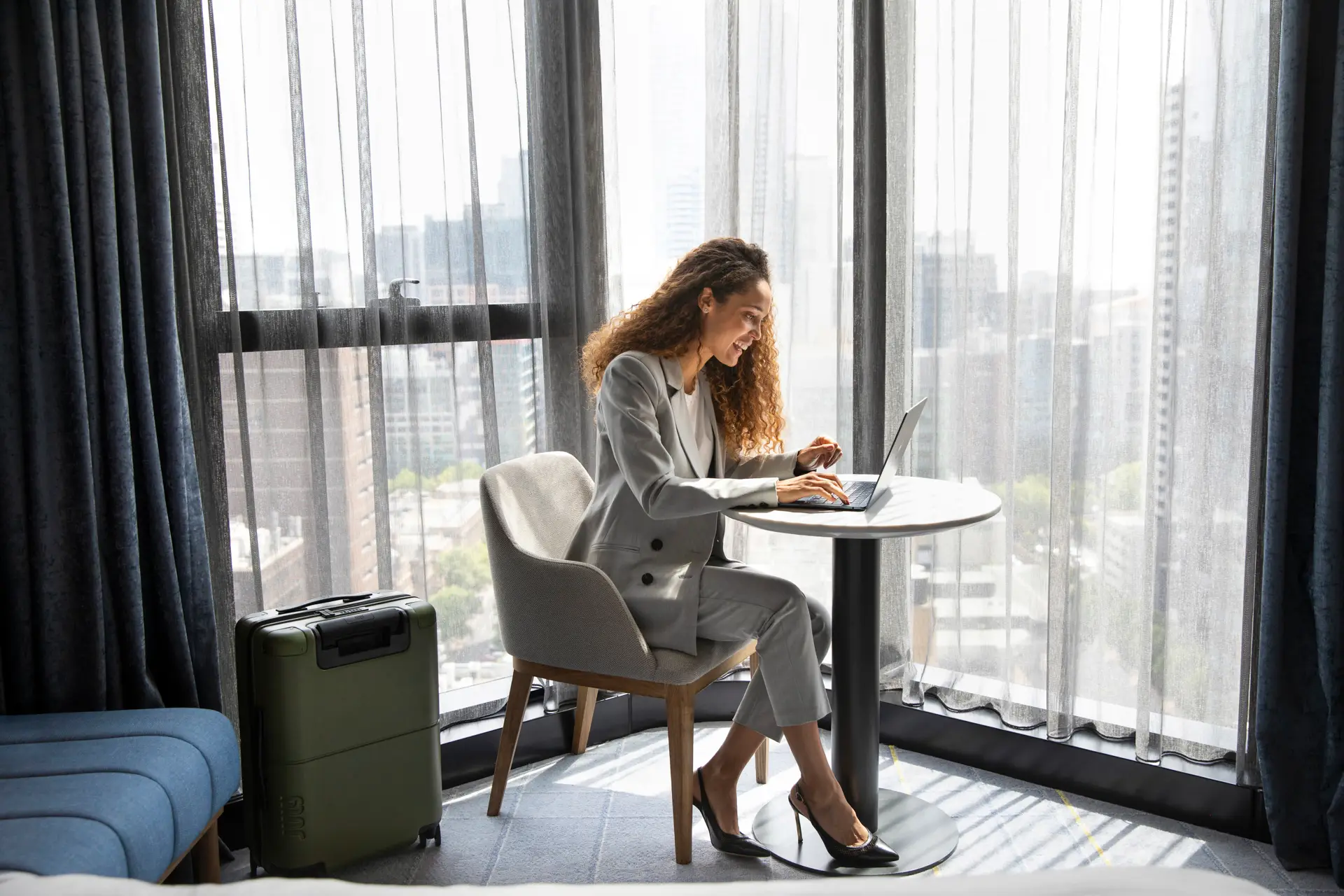 Guest working on a laptop at a round table beside a floor-to-ceiling window with city views
