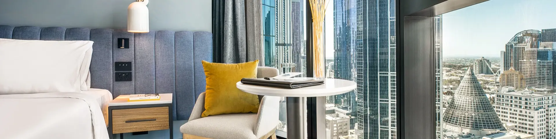 Guest room with bedside seating and a round table beside floor-to-ceiling windows overlooking the city