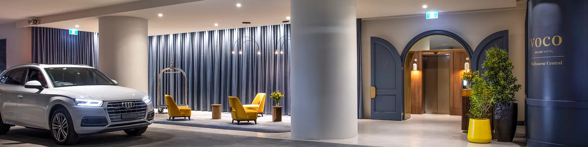 Hotel arrival area with a silver car, luggage trolley, lounge chairs and voco Melbourne Central signage