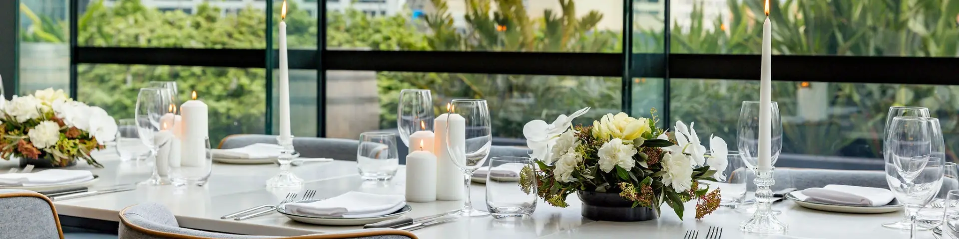 Event table set with candles, glassware, white napkins and floral arrangements beside large windows