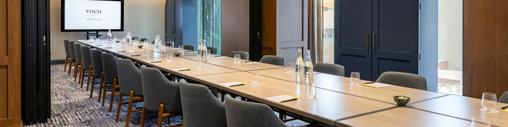 Boardroom with a long meeting table, grey chairs, water bottles and presentation screen