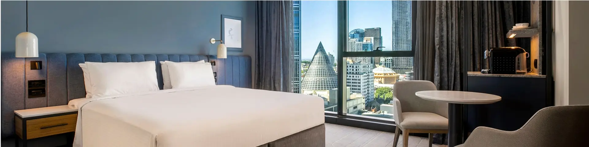 Guest room with a large bed, city views, round table, chairs and integrated coffee station