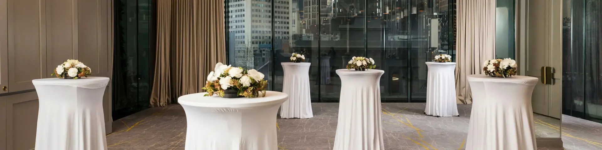 Cocktail event setup with tall white tables and floral arrangements beside floor-to-ceiling windows