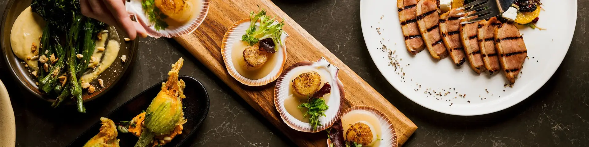 Shared dining spread with scallops, grilled meat, greens and a fried zucchini flower