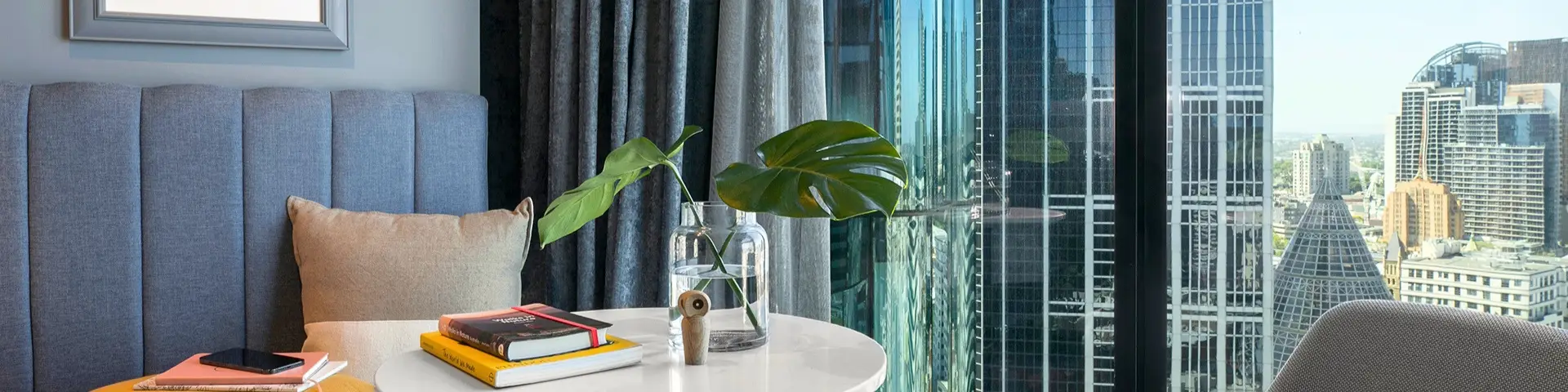 Blue window seat, round table and indoor greenery beside floor-to-ceiling windows overlooking the city