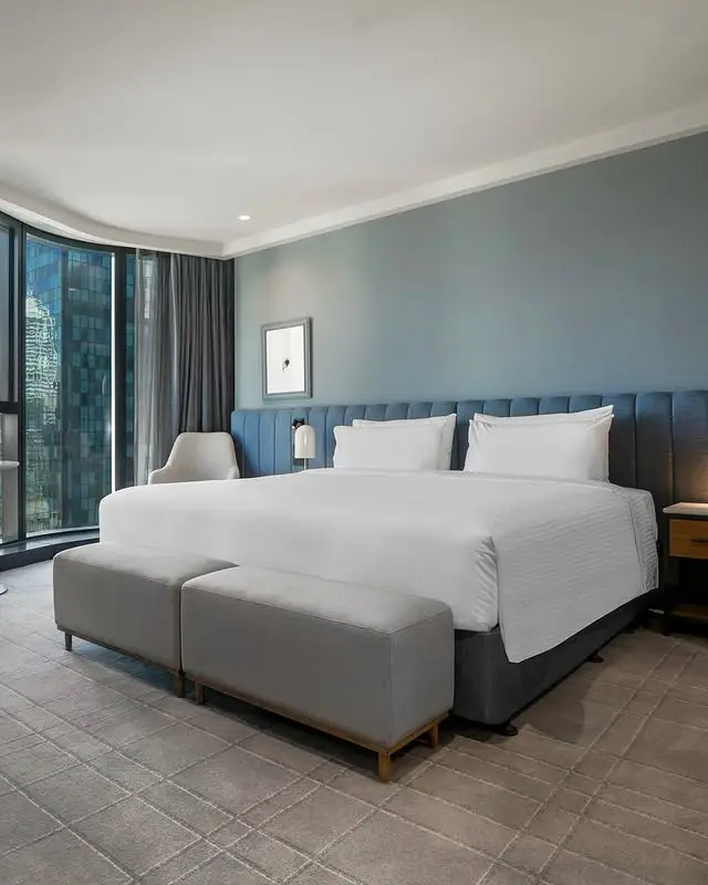 Spacious king room with white bedding, blue upholstered headboard and floor-to-ceiling windows at voco Melbourne Central